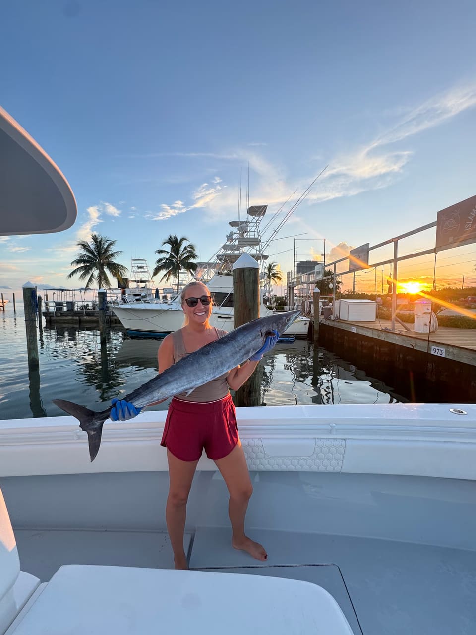 Smiling woman holding a large fish on a boat docked at sunset with palm trees and fishing boats in the background.