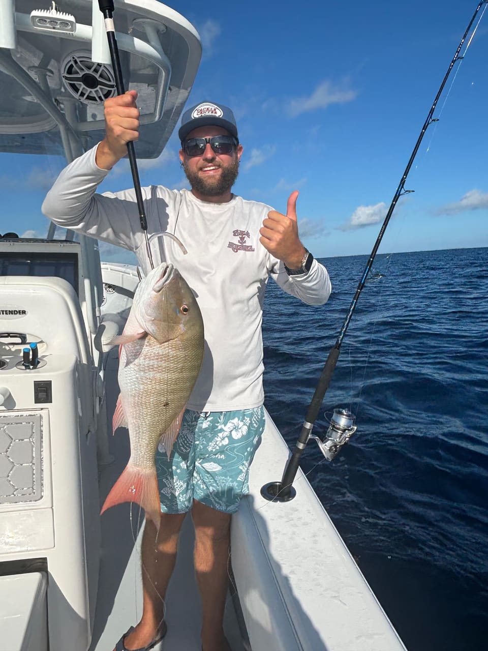 Angler giving a thumbs-up while holding a large mutton snapper on a charter boat in the open ocean, with fishing rods and reels visible behind him