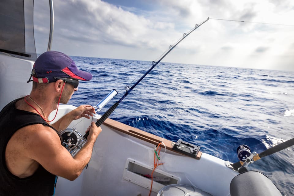 Angler rigging a trolling rod and reel on the stern of a sport fishing boat in the open ocean, with large swells and a cloudy sky behind him