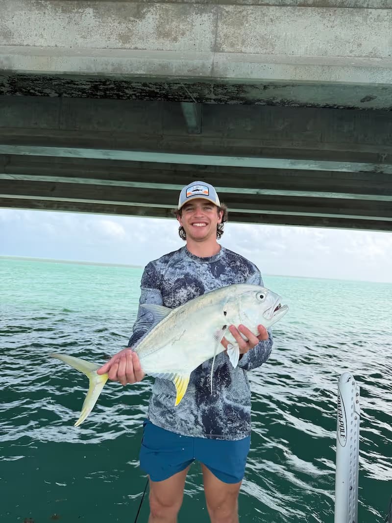 Smiling young man wearing a cap and camouflage shirt holding a large permit on a boat under a bridge with turquoise water in the background.